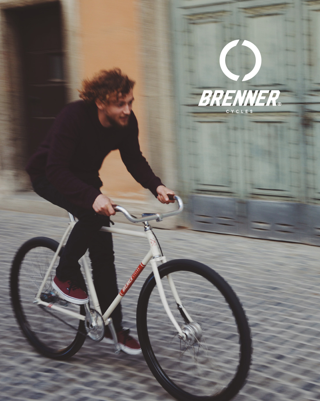 Ein Fahrer auf dem cremefarbenen Rad über Kopfsteinpflaster, oben rechts das Logo